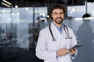 une professionnel Masculin médecin avec une stéthoscope des stands en toute confiance dans une hôpital paramètre, en portant une tablette et souriant. photo
