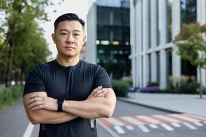 portrait de une sérieux Jeune asiatique des sports entraîneur homme permanent sur une ville rue, avec le sien bras franchi sur le sien poitrine et à la recherche en toute confiance à le caméra. photo