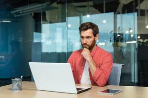 sérieux en pensant homme d'affaire proche en haut, homme dans rouge décontractée chemise travail à l'intérieur moderne bureau, mature patron travail avec portable à lieu de travail tandis que séance photo