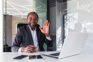 portrait de réussi mature africain américain chef, homme souriant et à la recherche à caméra séance à bureau à l'intérieur bureau, homme d'affaire agitant main geste de salutation et amitié. photo