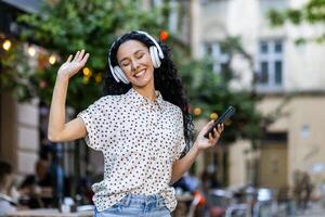 Jeune magnifique hispanique femme écoute à la musique chante le long de et danses tandis que en marchant dans soir ville, femme avec frisé cheveux les usages écouteurs et app sur téléphone. photo