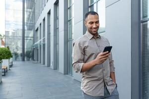 Latin américain homme d'affaire dans décontractée chemise en marchant près Bureau bâtiment de dehors, homme souriant et en utilisant téléphone, pigiste dactylographie message et navigation l'Internet pages photo