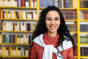 une Jeune hispanique femelle étudiant des stands dans de face de une coloré étagère à livres dans une bibliothèque, symbolisant éducation, connaissance, et diversité. photo