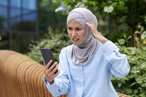 dérangé triste femme reçu en ligne notification message avec mal nouvelles, musulman femme dans hijab en portant téléphone, en utilisant application. photo