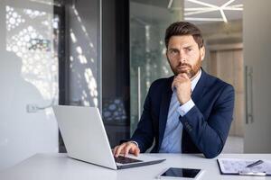 portrait de une sérieux Jeune homme d'affaire séance dans une costume à une bureau dans une moderne Bureau dans de face de une ordinateur portable, à la recherche en toute confiance à le côté tandis que en portant le sien main près le sien menton. photo