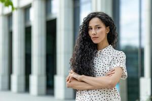 professionnel Jeune femme d'affaires avec frisé cheveux permanent en toute confiance à l'extérieur un bureau, bras franchi pendant une Pause. photo