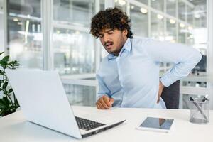 une Jeune Masculin Bureau ouvrier séance à le sien bureau dans une moderne bureau, expérience retour douleur tandis que travail sur le sien portable. photo