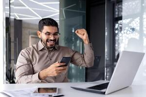 réussi homme d'affaire gagnant à l'intérieur Bureau à lieu de travail, homme reçu en ligne notification de la victoire gagner, ouvrier à bureau avec portable célébrer triomphe. photo