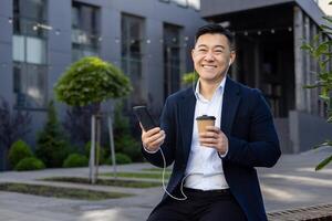 portrait de une Jeune asiatique homme séance dans une costume près le Bureau centre portant écouteurs en portant une tasse de café et une mobile téléphone, et à la recherche à le caméra avec une sourire. photo