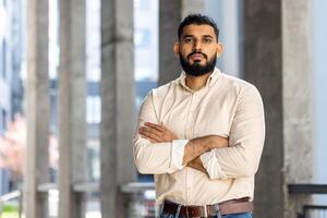 portrait de une Jeune musulman homme dans une chemise permanent à l'extérieur un Bureau bâtiment, traversée le sien bras plus de le sien poitrine et en toute confiance à la recherche à le caméra photo