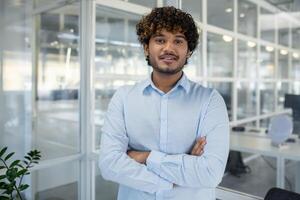 une portrait de une sur de soi Jeune homme d'affaire avec frisé cheveux, permanent bras franchi dans une rempli de lumière, contemporain Bureau environnement. photo