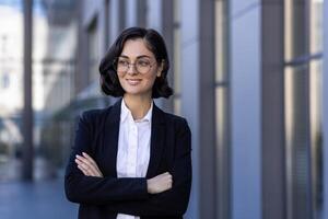 portrait de une Jeune affaires femme dans des lunettes et une costume permanent à l'extérieur un Bureau bâtiment avec sa bras franchi sur sa poitrine. il regards une façon avec une sourire, une fermer photo. photo