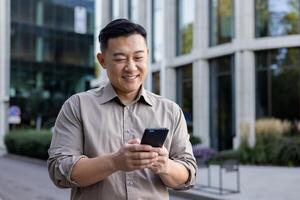 une souriant Jeune asiatique homme détient une mobile téléphone dans le sien mains et les types messages, des textes, appels. permanent souriant sur le ville rue. photo