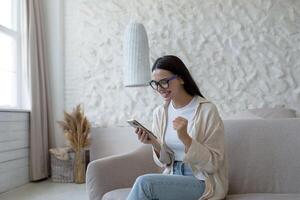 une Jeune content femme est séance à Accueil sur le canapé avec une téléphone. pièces en ligne Jeux, loterie, tombola photo