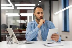 une professionnel Masculin entrepreneur dans une bleu chemise est absorbé dans en train de lire financier rapports sur une numérique tablette à le sien espace de travail, illustrant le concept de affaires analyse, numérique flux de travail, et La technologie dans le lieu de travail. photo