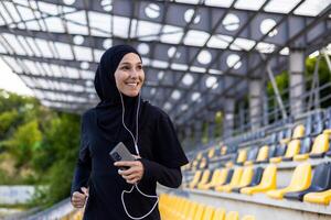 Jeune magnifique musulman femme fonctionnement dans hijab dans stade, sportive souriant dans écouteurs en portant téléphone, écoute à l'audio livre. photo