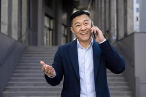 portrait de une souriant et réussi Jeune asiatique affaires homme dans une costume permanent à l'extérieur une bâtiment et parlant sur le téléphone. faire des gestes avec mains et à la recherche à caméra. photo