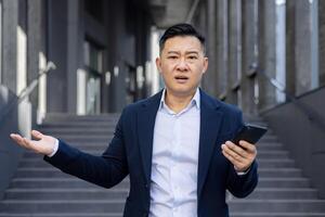 portrait de une déçu et sous le choc Jeune asiatique homme permanent à l'extérieur une affaires bâtiment, en portant une téléphone, à la recherche dérangé à le caméra et diffusion le sien mains. photo