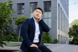 à l'extérieur le Bureau bâtiment, une Jeune asiatique homme dans une costume est séance sur une banc, en portant le sien main par le sien cou, fatigué de travail, Faire une massage, sentiment douleur dans le sien muscles. photo