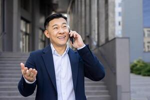 souriant asiatique homme d'affaire dans une costume fabrication une téléphone appel tandis que faire des gestes avec main en plein air, montrant la communication et succès. photo