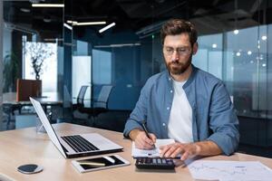 sérieux et concentré financier comptable sur papier travail à l'intérieur bureau, mature homme en utilisant calculatrice et portable pour calculateur rapports et résumant comptes, homme d'affaire à travail dans décontractée vêtements. photo