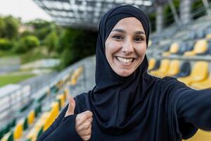 souriant femme dans une noir hijab capture une selfie avec une pouces vers le haut à un Extérieur des sports stade avec Jaune des places. photo