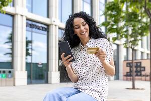 Jeune magnifique femme en marchant dans le ville, satisfait Latin américain femme avec frisé cheveux détient téléphone et banque crédit carte dans mains, fait du en ligne achats Heureusement livres prestations de service et choisit en ligne. photo