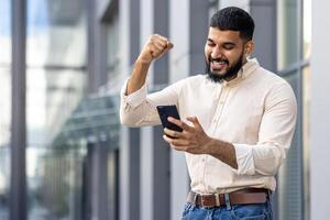 joyeux homme célébrer Succès avec téléphone intelligent dans Urbain réglage photo