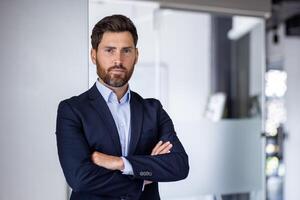 portrait de mature expérimenté financier homme d'affaire, homme en pensant sérieusement à la recherche à caméra avec franchi bras, sur de soi investisseur banquier à l'intérieur Bureau lieu de travail dans affaires costume. photo