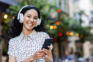 joyeux femme avec écouteurs en utilisant téléphone intelligent en plein air. content, connecté, Urbain la vie concept avec bokeh lumières Contexte. photo