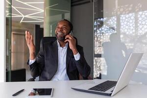 mature expérimenté patron parlant sur le téléphone à lieu de travail à l'intérieur le bureau, souriant homme d'affaire ayant une conversation avec les partenaires et collègues, satisfait avec le résultats de le négociation. photo