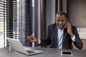 furieux diverse Masculin dans vêtements de cérémonie en portant lunettes tandis que ayant désagréable téléphone conversation. en colère loin ouvrier audition mal rupture nouvelles tandis que séance par bureau avec portable ordinateur. photo