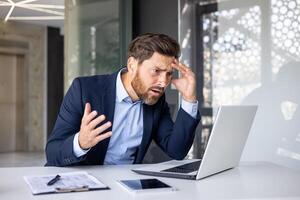 sous le choc et surpris Jeune homme d'affaire homme séance à bureau dans Bureau et à la recherche sérieusement à portable moniteur en portant diriger. photo