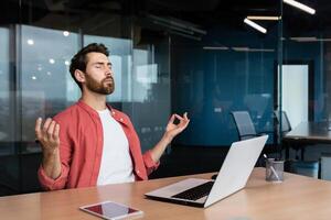 homme d'affaire dans une chemise repos et méditer dans le Bureau dans le lotus position, une homme à le lieu de travail se détend pendant le journée séance à le lieu de travail avec une portable avec le sien yeux fermé. photo