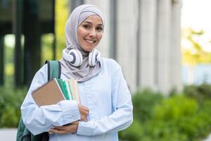 portrait de une de bonne humeur Jeune femme portant une hijab, en portant livres et portant écouteurs, permanent à l'extérieur avec une content expression. photo