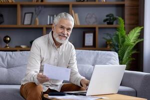 une de bonne humeur personnes âgées homme travaux de le sien confortable vivant chambre, en utilisant une portable et en portant documents. il sourit chaleureusement, exprimer contentement et productivité tandis que travail à distance. photo