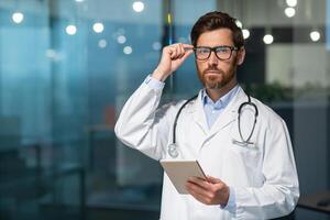 portrait de une mature médecin avec une barbe, le homme est à la recherche à le caméra dans des lunettes et en portant une tablette ordinateur dans le sien mains, le médecin est travail à l'intérieur le Bureau de une moderne clinique. photo