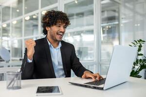 une joyeux Jeune homme est assis dans une moderne bureau, célébrer une réussi moment avec le sien main élevé dans la victoire tandis que en utilisant une portable. photo