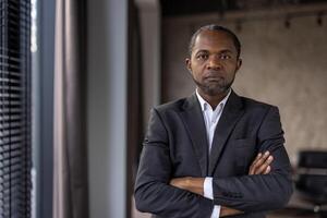 professionnel africain homme dans costume des stands avec bras franchi dans un bureau, exsudant confiance et direction. photo