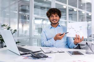 portrait de Jeune réussi financier formalités administratives à l'intérieur bureau, homme souriant et à la recherche à caméra spectacles graphique avec positif dynamique de économique bénéfices, homme d'affaire à travail avec portable. photo