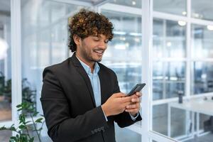 réussi financier investisseur travaux à l'intérieur Bureau à travail, homme d'affaire dans affaires costume les usages Téléphone près fenêtre, homme sourit et lit bien nouvelles en ligne de téléphone intelligent. photo