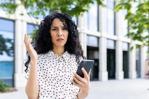 agacé Indien femme permanent sur le rue avec une téléphone dans sa mains, triste de mal nouvelles, indigné par une défectueux téléphone intelligent, à la recherche à le caméra. photo