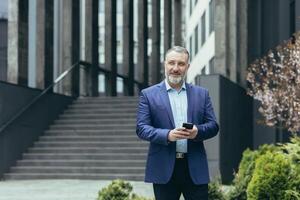 un plus âgée aux cheveux gris homme, une homme d'affaire dans une costume, est en voyageant à un autre ville, à un autre pays. il est en portant le téléphone dans le sien mains, à la recherche pour directions sur GPS. des promenades près une moderne bâtiment. photo