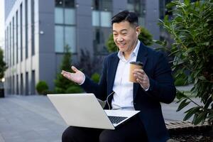 souriant asiatique Masculin Bureau ouvrier séance près bâtiment sur banc portant écouteurs, en buvant café et bavardage sur appel de portable. photo