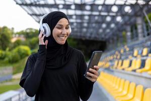 musulman sportive dans hijab à stade avec écouteurs et téléphone dans mains, femme pendant actif exercice et aptitude, en plein air écoute à en ligne la musique de l'audio livres et podcasts application. photo