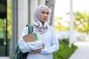 portrait de une concentré musulman femme avec hijab, écouteurs, et manuels faire un pas à l'extérieur dans un académique paramètre. photo