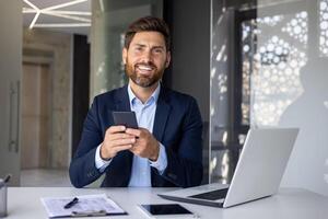 portrait de une Jeune homme d'affaire séance dans une costume à une bureau à l'intérieur le bureau, en portant une mobile téléphone, souriant et à la recherche à le caméra. photo