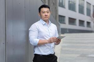 portrait de une Jeune Masculin asiatique étudiant permanent sur le rue sur le étudiant Campus. détient une tablette dans le sien mains, les types, études, prépare pour examens. il regards sérieusement dans le caméra. photo