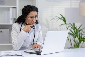 latino femme médecin en utilisant ordinateur portable, à la recherche perplexe ou concerné, dans une médical Bureau paramètre. photo