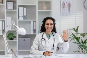 amical latina médecin dans une blanc manteau salutation une patient pendant une virtuel consultation dans sa bureau. photo